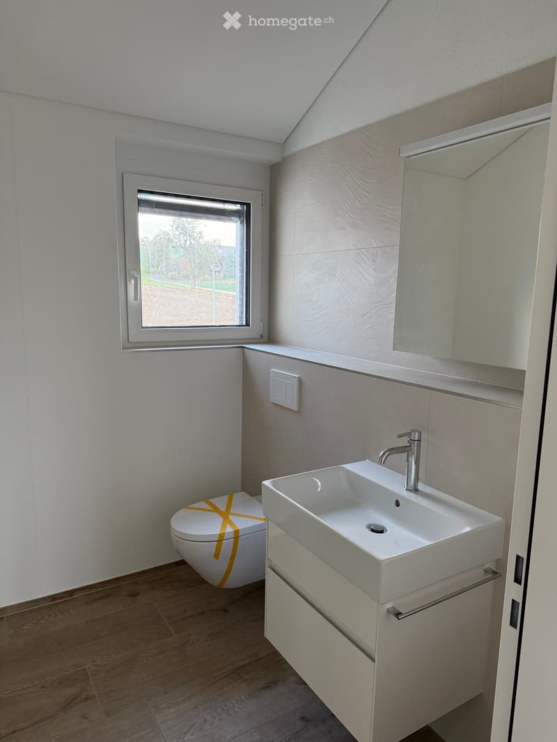 Modern bathroom with a white sink, chrome faucet, toilet, and large mirror above the sink. The room features light-colored tiles and wooden flooring.