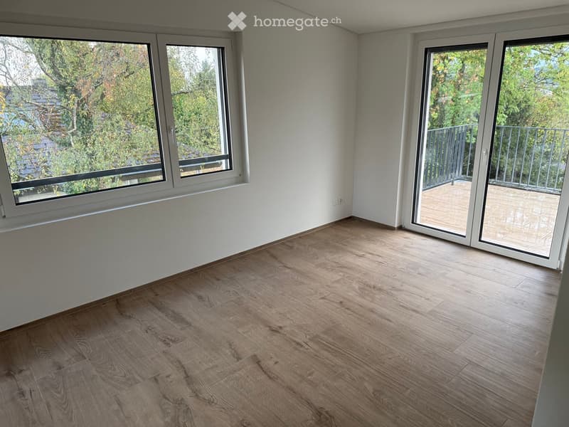 Empty room with wooden flooring, large windows, and sliding doors leading to a balcony.