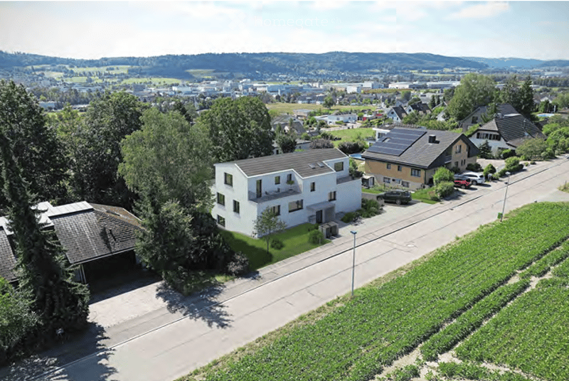 Aerial view of a modern two-story house with a white facade, grey roof, and surrounding greenery. The house features a garage and parking area, with neighboring houses and a scenic landscape in the background.