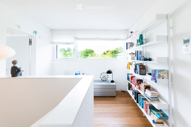 Bright, minimalist room with white walls, wooden floor, large window with blinds, built-in bookshelf filled with books and decorative items, small table with a lamp and sculpture, and a chair in the corner.