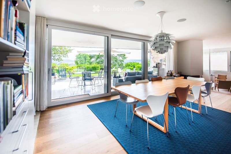 Bright dining area with a large wooden table, modern chairs, a blue rug, and a view of a garden through large glass doors.