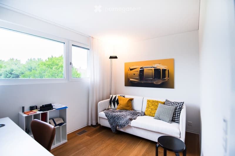 Modern living room with white walls, wooden flooring, large windows, a white sofa with colorful pillows, a black stool, a small table, and a shelf with books and decorative items.