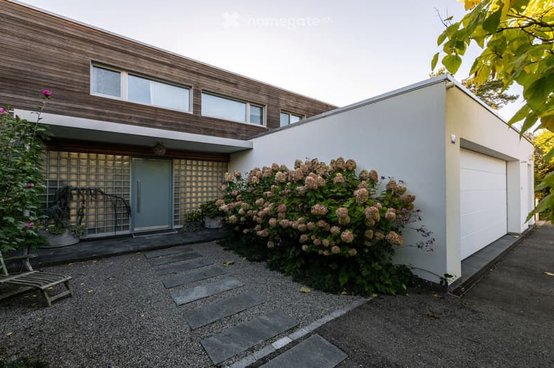Modern two-story house with wooden facade, white garage, and landscaped garden area with shrubs and a bench.