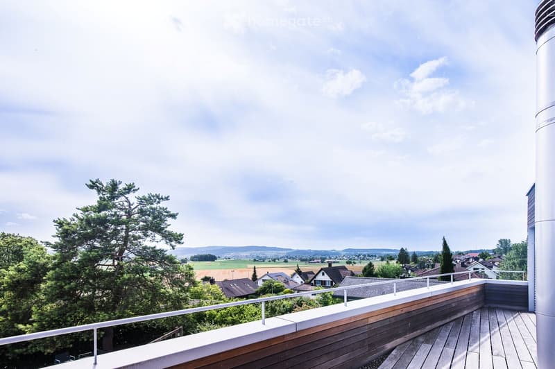 Wooden balcony with white railing, overlooking a rural landscape with trees, fields, and distant houses.