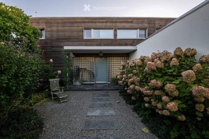 Modern house exterior with wooden facade, large windows, and a small garden area with a bench, shrubs, and flowering plants.