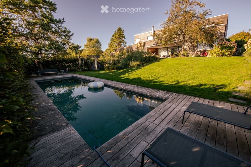 A modern outdoor area with a swimming pool, wooden decking, lounge chairs, and a well-maintained lawn surrounded by trees and shrubs.