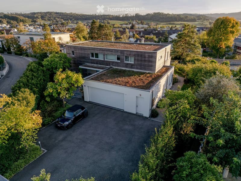 Modern two-story house with a green roof, garage, and parking area surrounded by trees and greenery.