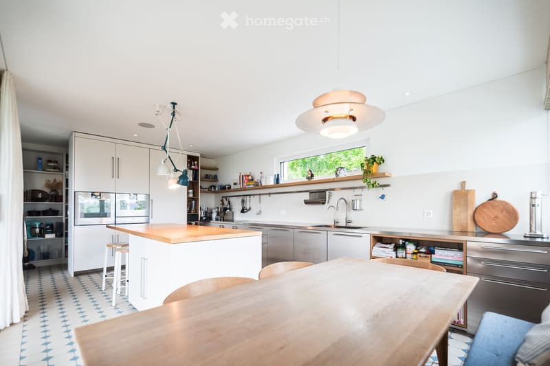 Modern kitchen with white cabinets, a central island, a dining table, and pendant lighting. Features stainless steel appliances, wooden countertops, and a mix of open shelving and built-in storage.