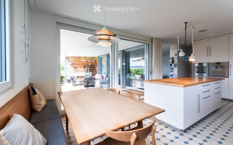 Bright, modern kitchen and dining area with white cabinets, wooden island, pendant lights, and a large window opening to a patio. Includes a wooden dining table, chairs, and a bench with cushions.