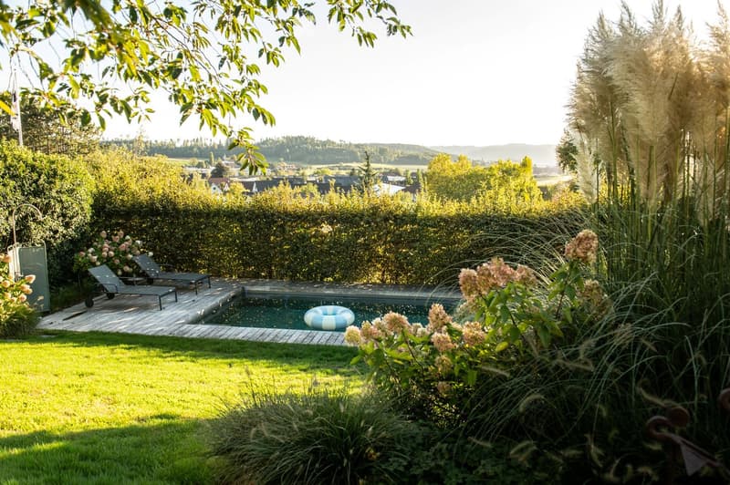 A well-maintained garden with a swimming pool, lounge chairs, and lush greenery, featuring a scenic view of a countryside landscape.