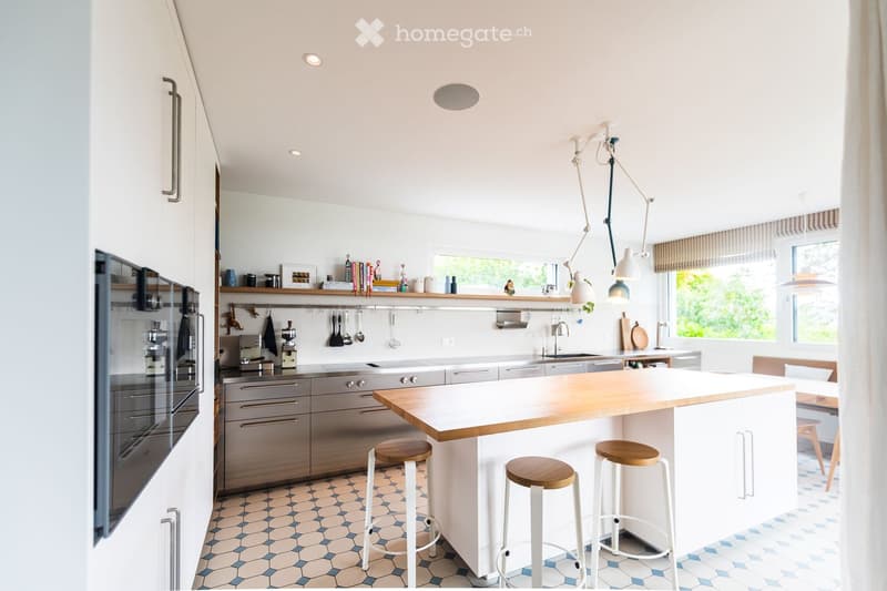 Modern kitchen with white cabinets, stainless steel appliances, a central island with wooden countertop, and four white stools. The space features pendant lights, open shelving with books and decorative items, and a sleek design.