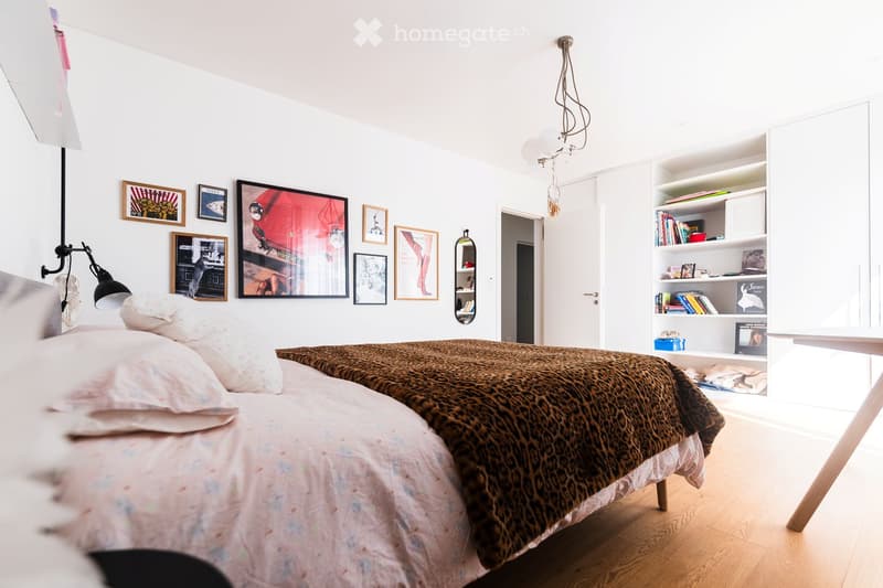Modern bedroom with a bed, leopard print throw, white bedding, built-in shelves with books and decorative items, framed artwork, and a mirror on the wall.
