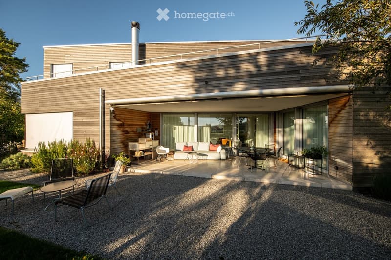 Modern house with a spacious outdoor patio area featuring a sofa, tables, and chairs. The patio is connected to the house via large glass doors, and the surrounding area includes gravel pathways and greenery.