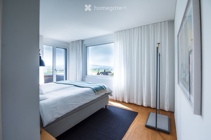 Modern bedroom with a large bed, white bedding, a blue throw, wooden flooring, large windows with white curtains, and a coat rack.