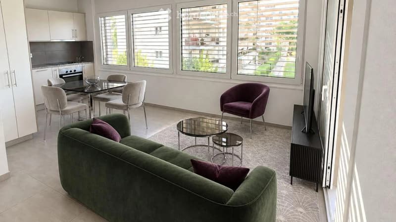 Modern living room with a green sofa, coffee table, glass dining table, chairs, and a TV stand. The room features large windows with blinds and a view of the building exterior.