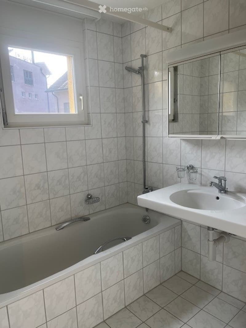 Bathroom with white tiled walls and floor, a bathtub, a sink with a mirror above it, and a showerhead on the wall.