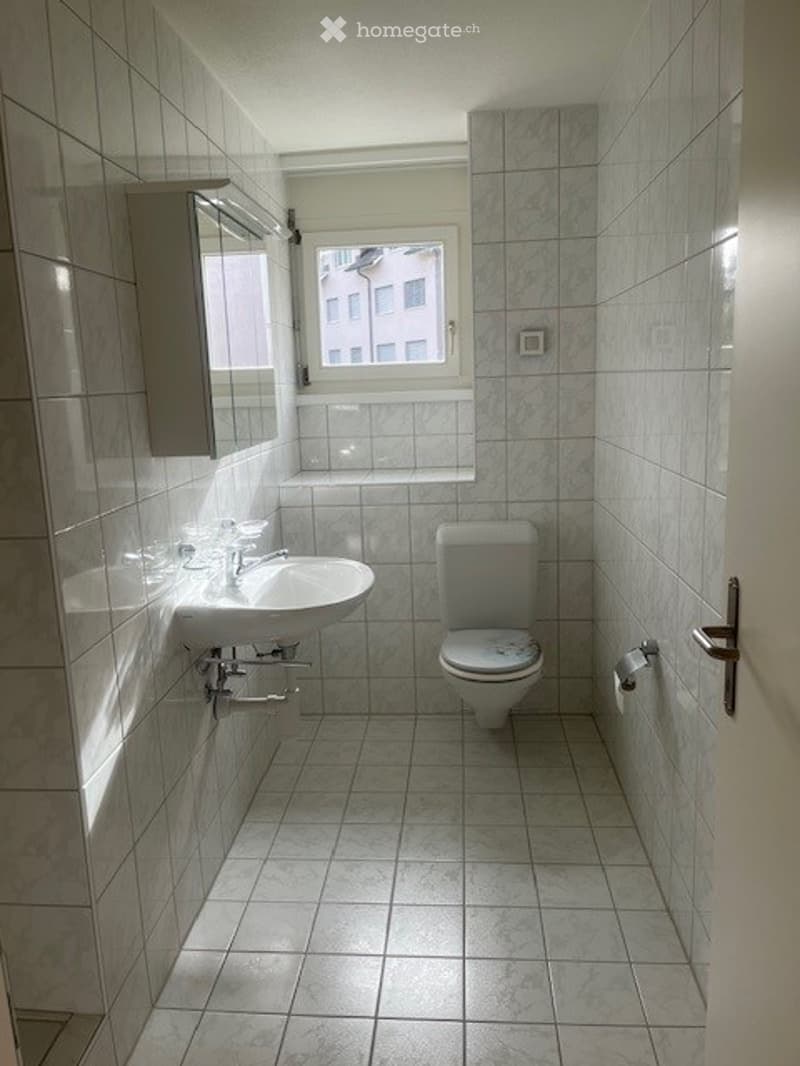 Modern bathroom with white tiled walls and floor, a sink with a faucet, a toilet, a mirror, and a small cabinet.
