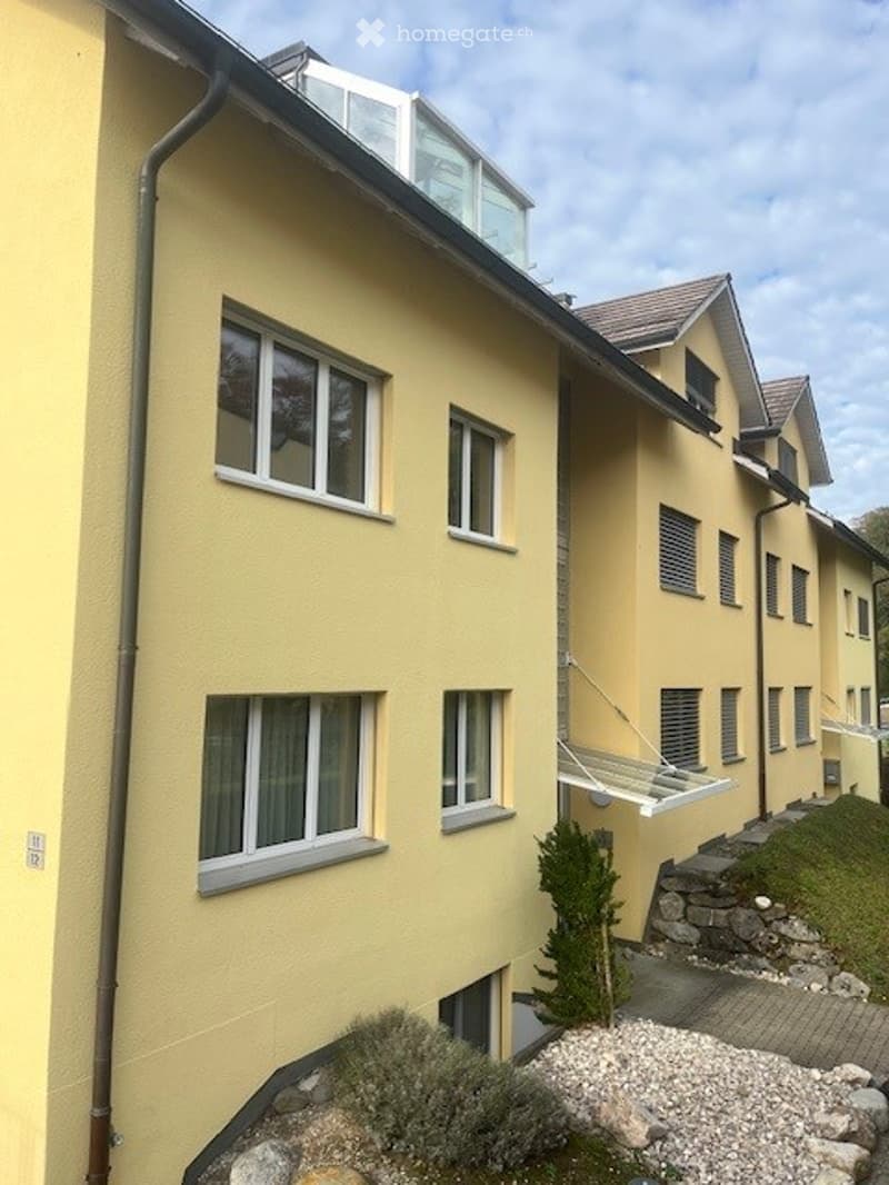 A modern yellow apartment building with multiple windows, a small balcony, and a landscaped garden area with shrubs and gravel.