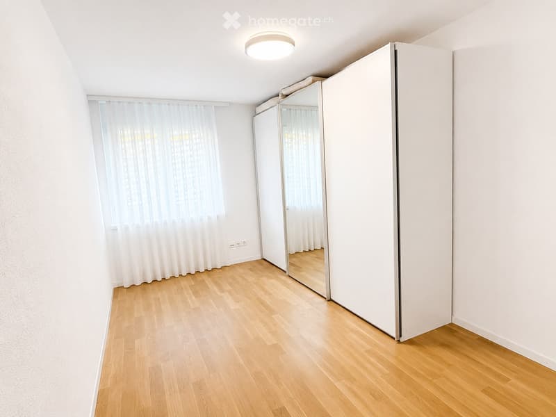 Empty room with light wooden flooring, white walls, large window with sheer curtains, mirrored sliding doors, and a ceiling light fixture.