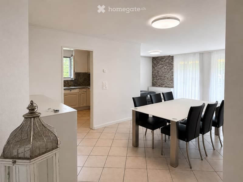 Modern dining area with a white dining table and black chairs, adjacent to a kitchen with light-colored cabinets and a window, featuring a decorative lantern on a side table.