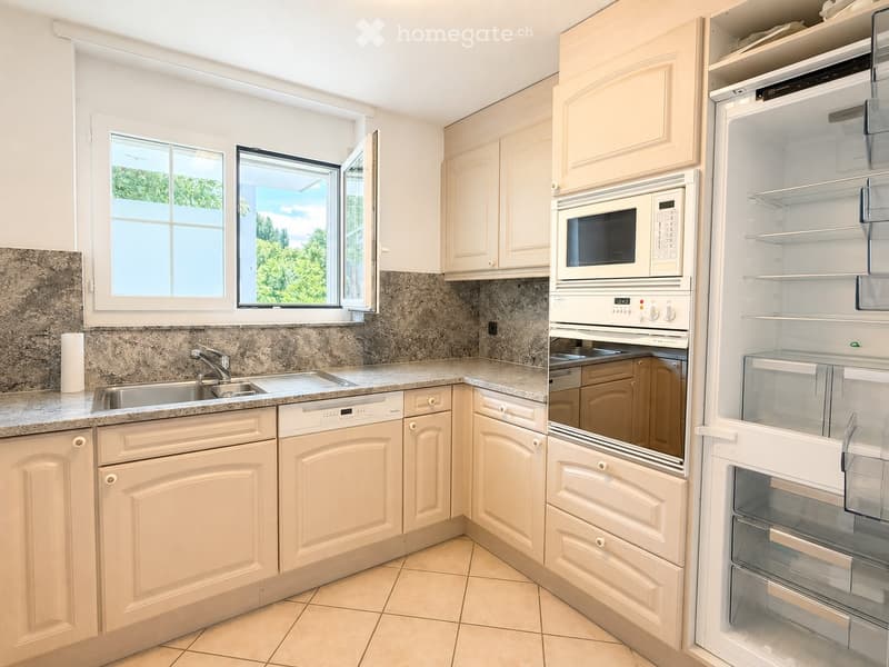 Bright kitchen with light-colored cabinets, granite countertops, and stainless steel appliances including a microwave, oven, refrigerator, and dishwasher. A window with a view of greenery is visible.