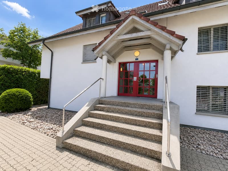 Two-story house with a red front door, white exterior, and a small porch with a roof. Stairs lead up to the entrance, flanked by metal railings. The house has multiple windows and a small garden area with trimmed bushes.