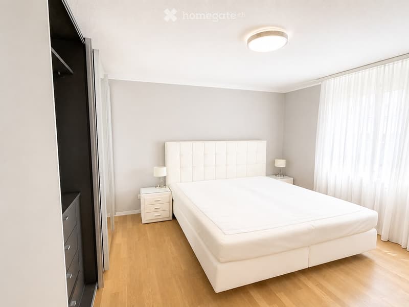 Modern bedroom with a large white bed, tufted headboard, two nightstands with lamps, and a tall wardrobe with mirrored doors.