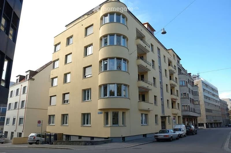 Multi-story residential building with light yellow facade, multiple windows, and balconies. Several cars are parked along the street in front of the building.