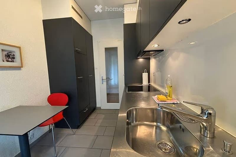 Modern kitchen with stainless steel sink, faucet, dark cabinets, and a small dining area with a table and red chair.