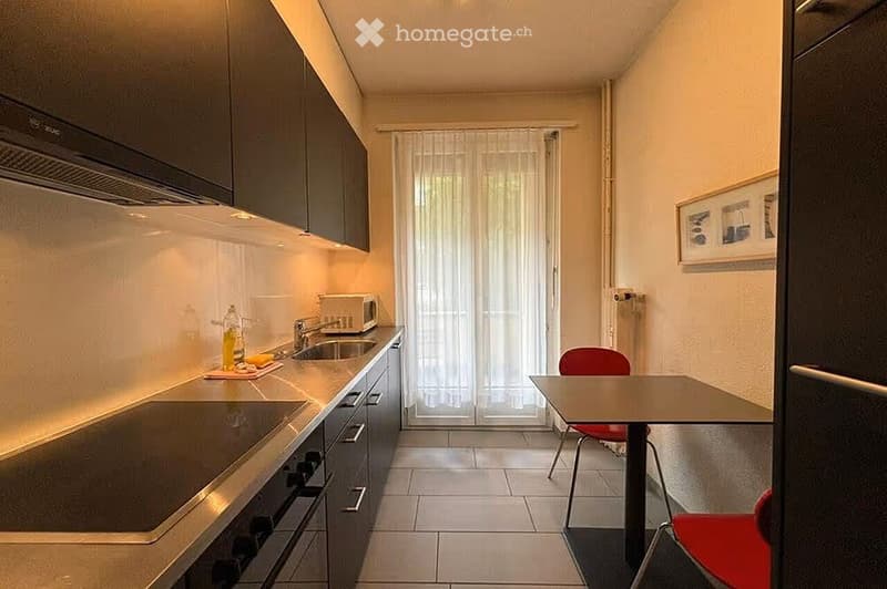 Modern kitchen with stainless steel appliances, a small dining area, and a window with sheer curtains.