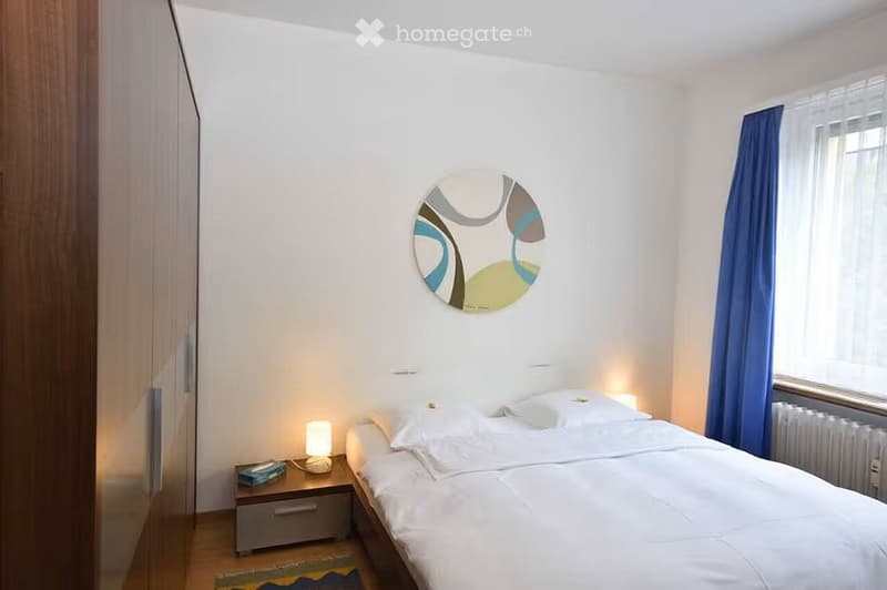 Modern bedroom with a double bed, nightstand, lamp, and a decorative wall mirror. Blue curtains cover the window, and a small rug is placed near the bed.
