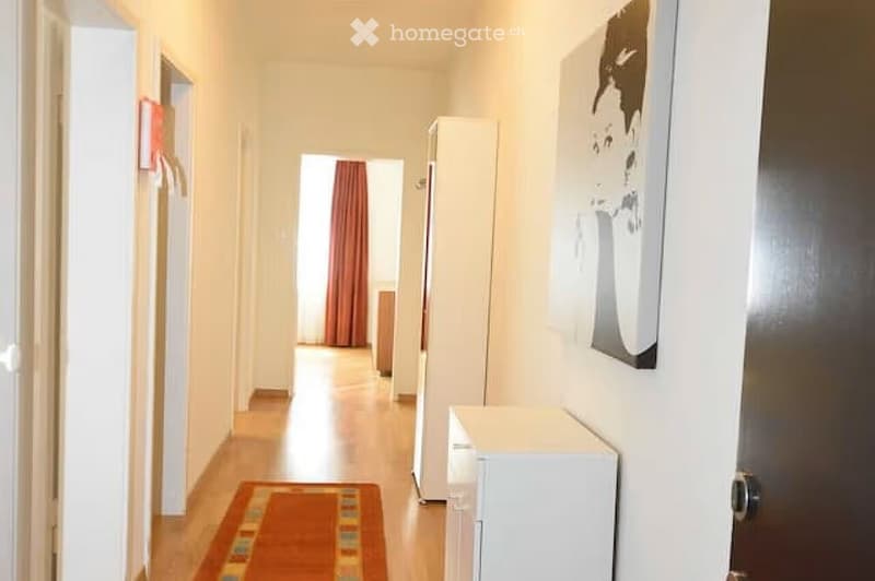 A narrow hallway with wooden flooring, white walls, and a small rug. A refrigerator is visible on the right side, and a doorway leads to another room with red curtains.