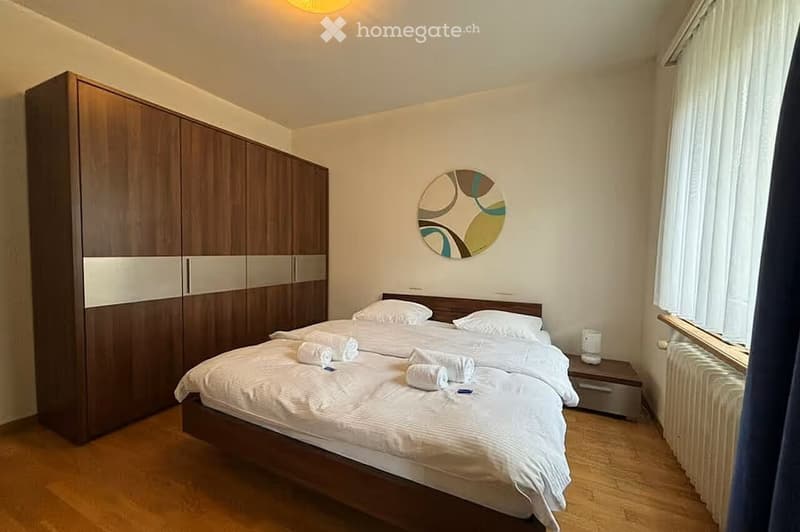 Cozy bedroom with a double bed, white linens, a large wooden wardrobe, a nightstand with a lamp, and a decorative wall mirror.