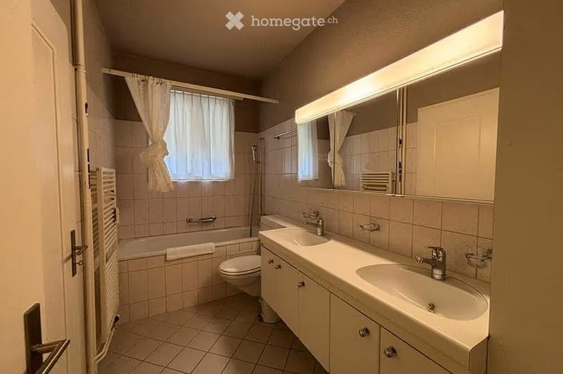 Bathroom with double sink, toilet, bathtub, large mirror, and tiled walls and floor.