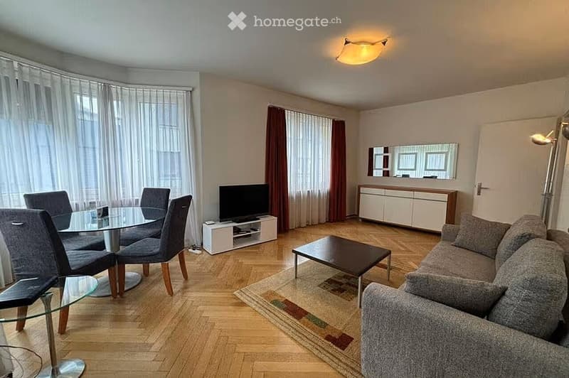 Modern living and dining area with a grey sofa, coffee table, dining table with chairs, TV, and a cabinet with a mirror above it. The space features wooden flooring, large windows with curtains, and a ceiling light fixture.