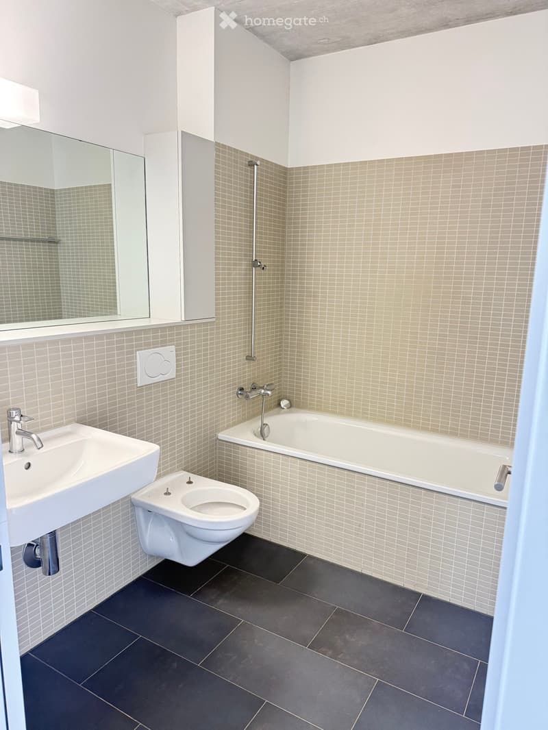 Modern bathroom with a white sink, toilet, bathtub, and mirror. Beige tiled walls and dark grey tiled floor.