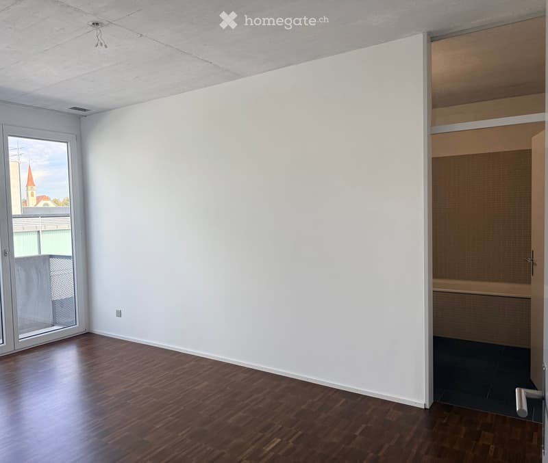 Empty room with wooden flooring, white walls, and large windows offering a view of a church spire. No furniture or equipment is present.