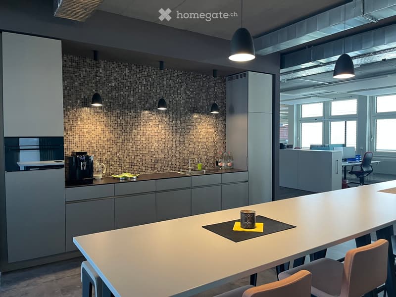 Modern kitchen area with grey cabinets, a mosaic tile backsplash, and a long white table with chairs. Coffee machine, sink, and various small items are visible.