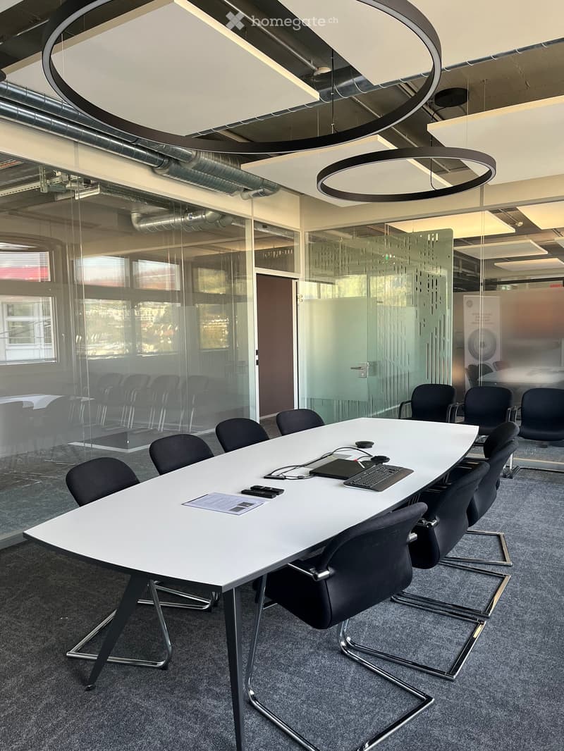 Modern conference room with a white rectangular table surrounded by black chairs, a laptop, keyboard, and remote controls on the table, with glass partitions and a view of another room.