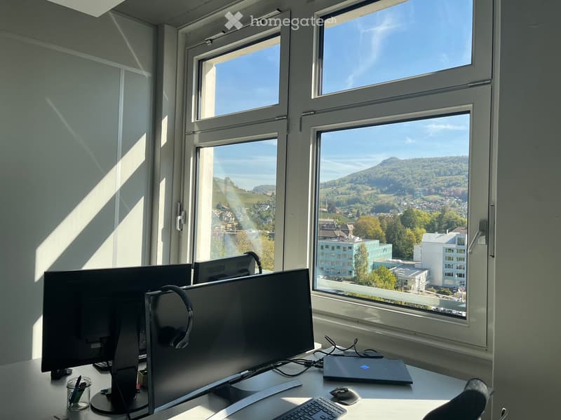 Office space with large windows offering a scenic view, equipped with a laptop, keyboard, mouse, headphones, and a screen.