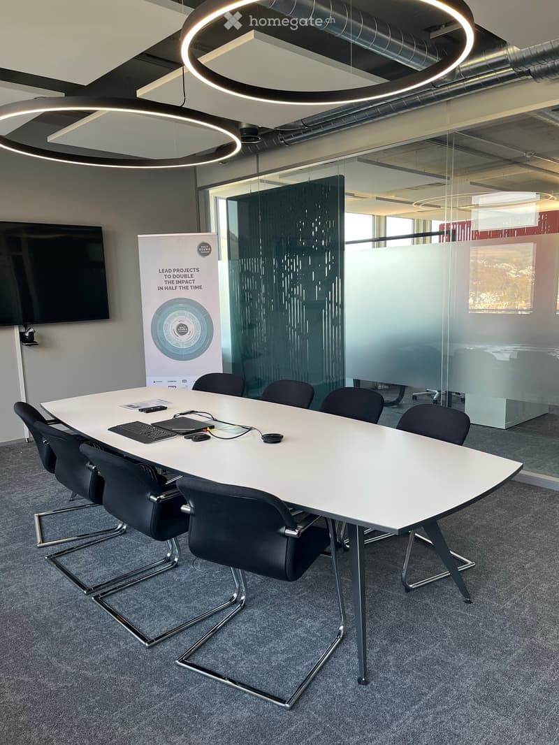 Modern conference room with a long white table, black chairs, laptop, and a large screen. A poster is displayed on the wall, and the room features large windows and modern lighting.