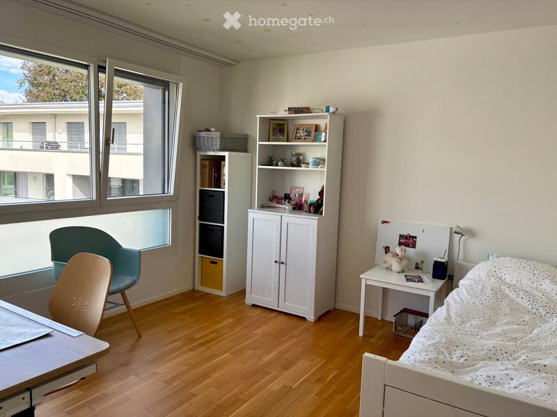 A well-lit bedroom with wooden flooring, a bed with a patterned blanket, a white cabinet, a shelf with decorative items, a small table with a lamp, and a couple of chairs near the window.