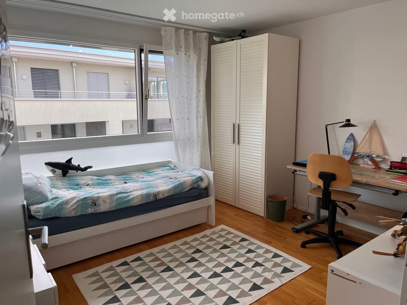 A well-lit bedroom with a single bed, desk, chair, wardrobe, and decorative items including a toy shark and model sailboat.