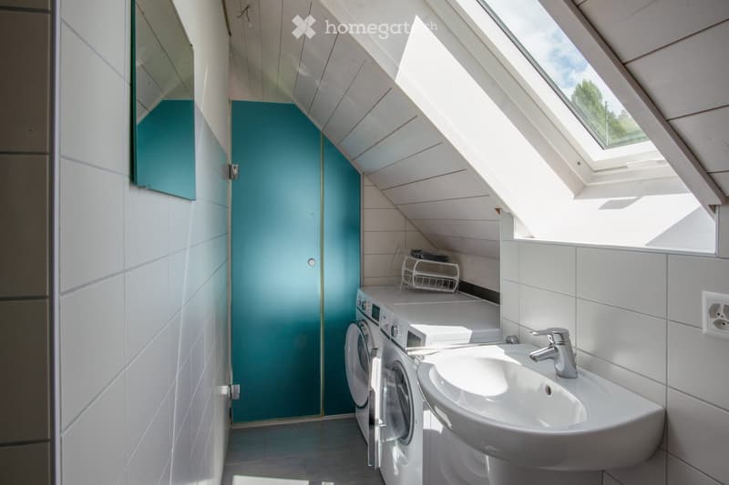 Modern bathroom with white tiles, a sink, washing machine, and a shower door. The room features a skylight and a small shelf with folded towels.