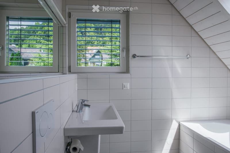 Modern bathroom with white tiled walls and floor, a single sink with a chrome faucet, a bathtub, and a window with blinds offering a view of greenery outside.