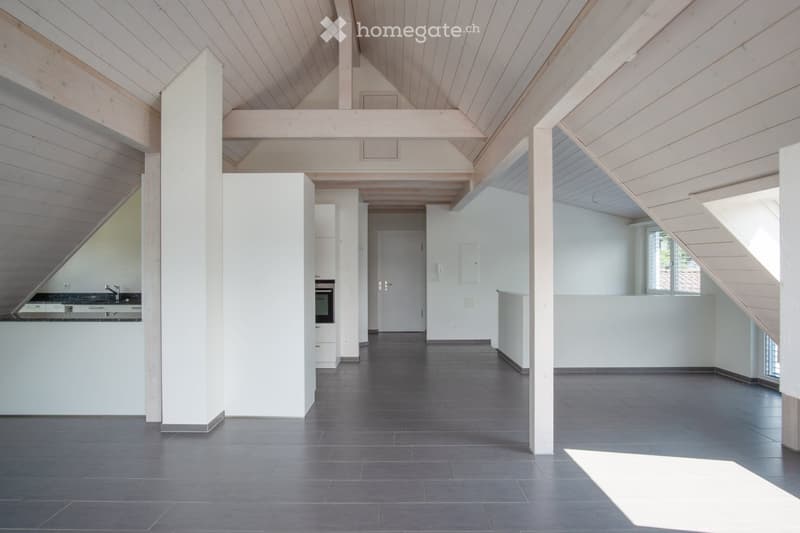 Open, modern loft-style space with exposed wooden beams and white walls, featuring a kitchen area with a sink and oven, and large windows allowing natural light.