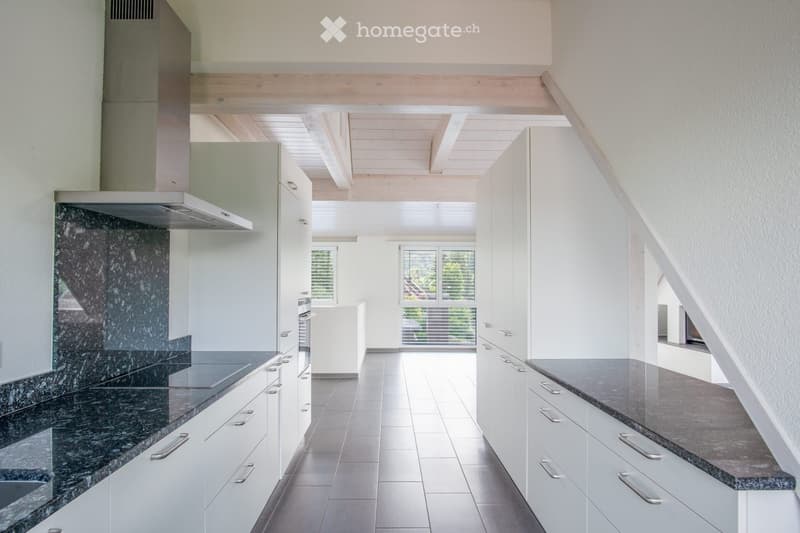 Modern kitchen with white cabinets, granite countertops, and a stainless steel range hood. The space features large windows and a high ceiling with exposed wooden beams.