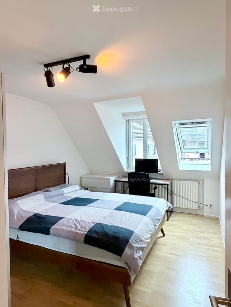 Cozy attic bedroom with wooden bed, desk, chair, and computer. Slanted ceilings and large windows provide natural light.