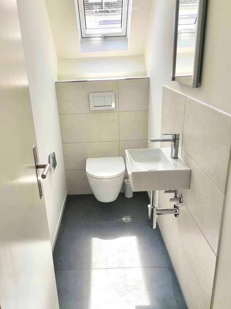 Modern bathroom with a toilet, sink, and mirror. Light-colored tiles and a dark floor.