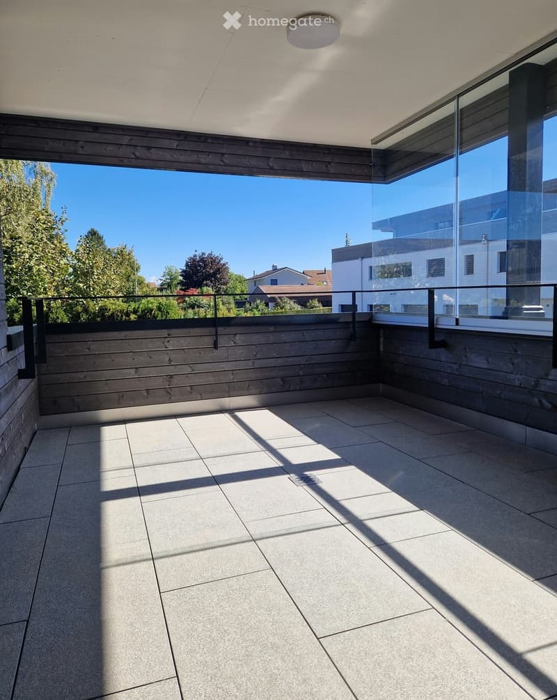 Modern balcony with tiled floor, wooden railing, and large glass windows offering a view of the surrounding area.